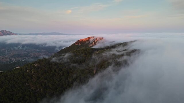 Aerial drone video of a forested mountain peak in Spain, bathed in warm sunlight, emerging above dense clouds at sunrise.