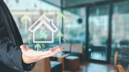 Businessman's hand presents a glowing house icon with rising percentage arrows, symbolizing real estate investment growth and increasing mortgage interest rates on a blurred office background.