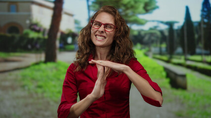 Woman making timeout gesture in outdoor park setting wearing red shirt and glasses with trees and path in background.