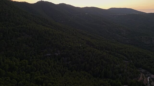 Aerial drone video of a forest-covered mountain peak in Spain at sunrise, with warm sunlight casting golden tones on rocky slopes.