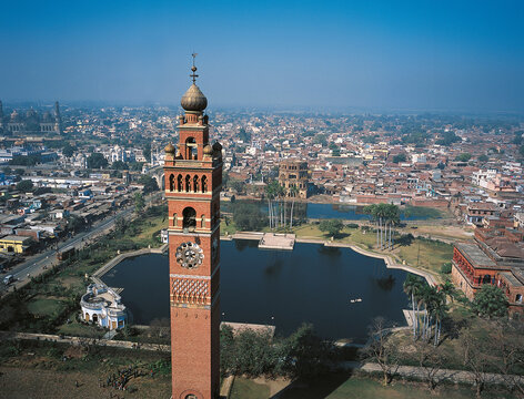 Aerial view of the iconic Hussainabad Clock Tower standing tall beside a tranquil pond, contrasting with the city's bustling rooftops, Lucknow, Uttar Pradesh, India.