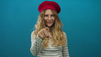 Naklejka premium Portrait of a woman whose hand is pointing at the camera in a studio as a blonde young subject wearing a beret and glasses against a blue backdrop flashes a smile.