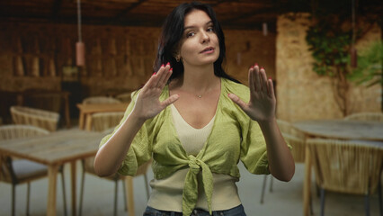 Young hispanic woman making a calm stop gesture with both hands in a restaurant building outdoors; serenity reassurance.