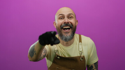 Obraz premium Bald man with beard laughing and pointing wearing apron and glove against pink background in an isolated setting displaying humor and joy with expressive gesture and tattoos.
