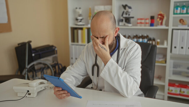 Bald man with a beard in a clinic examining a tablet, surrounded by medical equipment inside a hospital room.