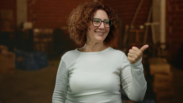Woman smiling indoors at construction site wearing glasses and a white sweater, showcasing a cheerful demeanor with curly hair against a blurred industrial background.