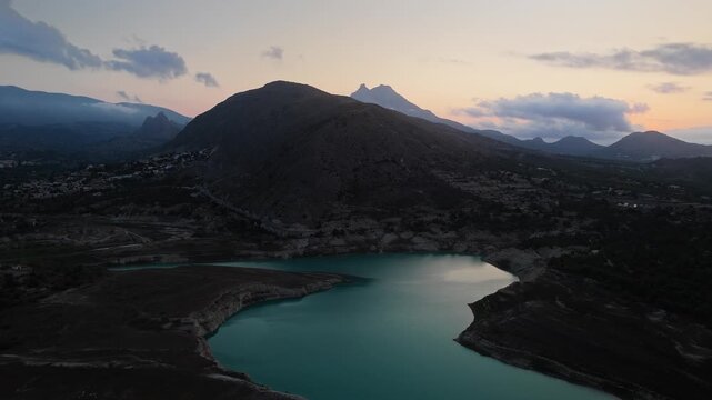 Drone video of a mountain lake in Spain at sunset, with calm turquoise water, rugged hills, and a dramatic sky blending warm and cool tones.