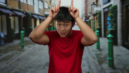Obraz premium Young man in red shirt making a horns gesture while standing in a lively urban street setting, surrounded by colorful buildings and a cheerful atmosphere.