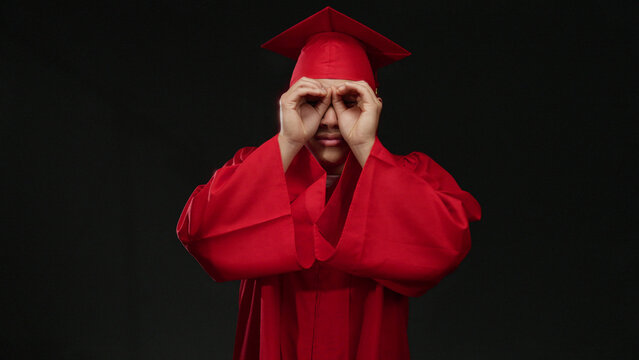 Young chinese man in red graduation cap and gown makes binocular gesture isolated on a black background, symbolizing curiosity and future vision.