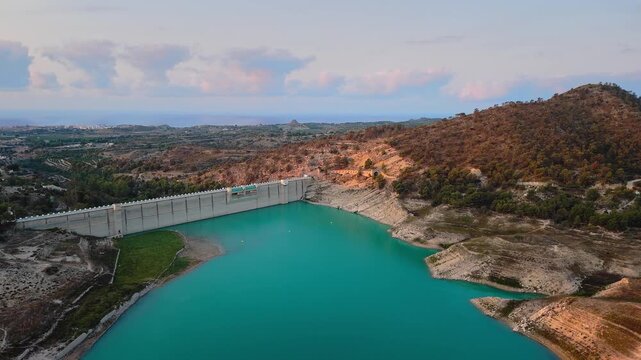 Drone video of a turquoise reservoir in Spain, showing the dam structure surrounded by hills and countryside under a soft pastel evening sky.