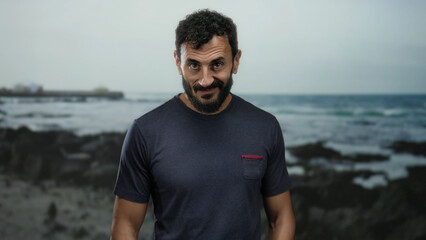 Hispanic man with beard standing confidently on rocky beach with ocean waves in background under cloudy sky wearing casual shirt during daytime.