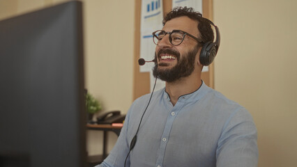 Hispanic bearded man wearing headset smiling during online meeting in a bright office, showcasing technology and communication in professional indoor workspace.