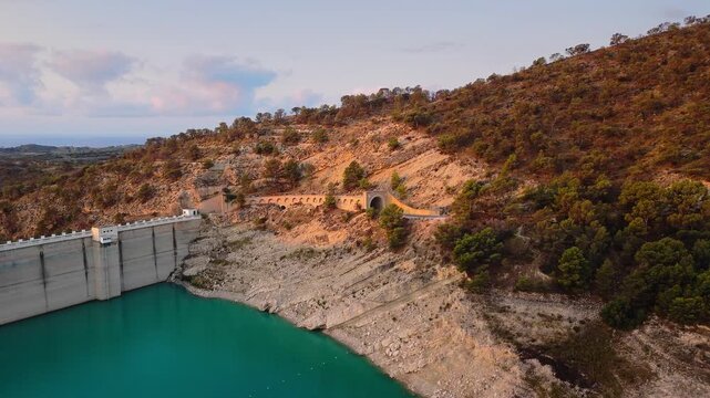 Drone video of a turquoise reservoir in Spain, showing the dam structure surrounded by hills and countryside under a soft pastel evening sky.