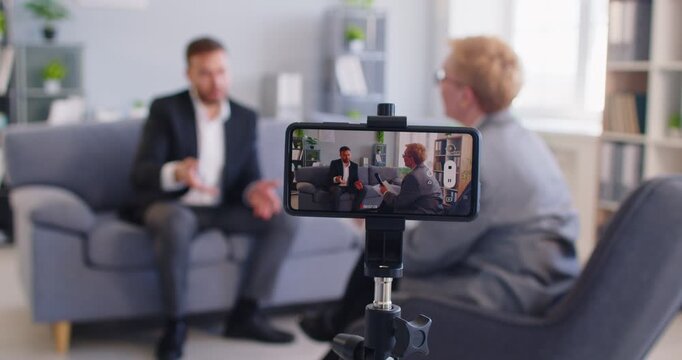 Close up of smartphone camera on tripod recording marketing podcast vlog or professional business video of two people. Businessman talking in TV show studio, giving interview to female journalist. - Powered by Adobe