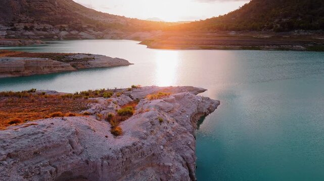 Drone video capturing the rocky lake shore in Spain at sunset, with golden sunlight reflecting on turquoise water and surrounding rugged terrain.