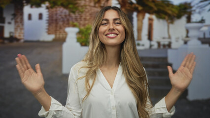Fototapeta premium Young blonde woman wearing white blouse showing hand size gesture in a sunny courtyard; confidence joy.