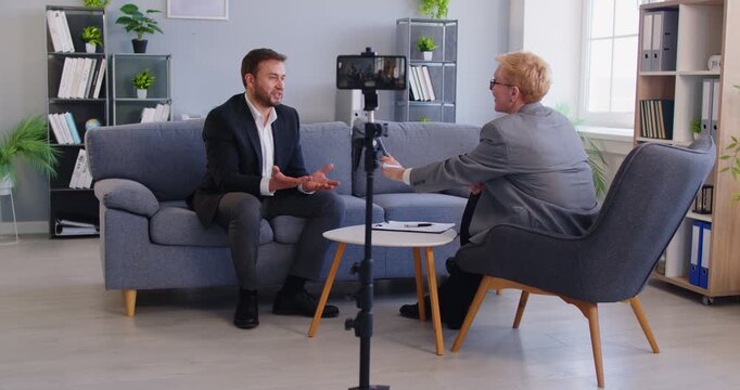 Young male company CEO speaker sitting on sofa in office workplace setting discusses business, work, finance questions with female media program host, phone on tripod recording digital interview video
