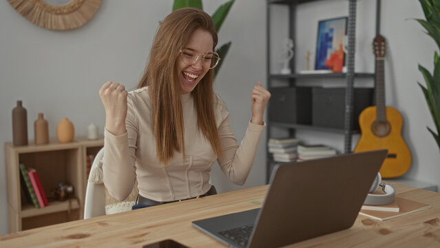 Woman pumping fists and smiling face at laptop on wooden desk in office; success celebration achievement triumph.