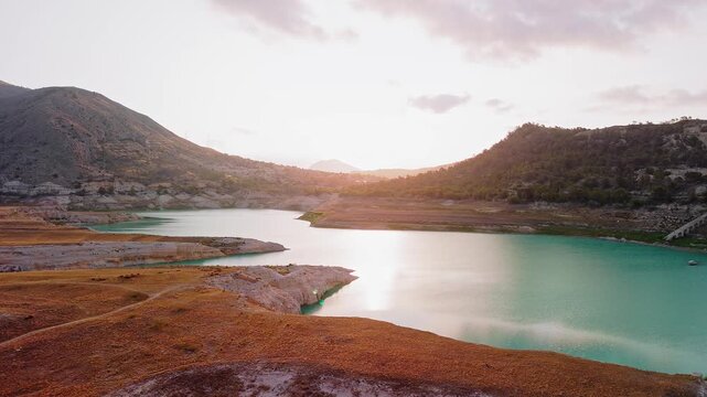 Drone video capturing a serene mountain lake in Spain at sunset, with golden light reflecting on turquoise waters and rugged hills in the background.