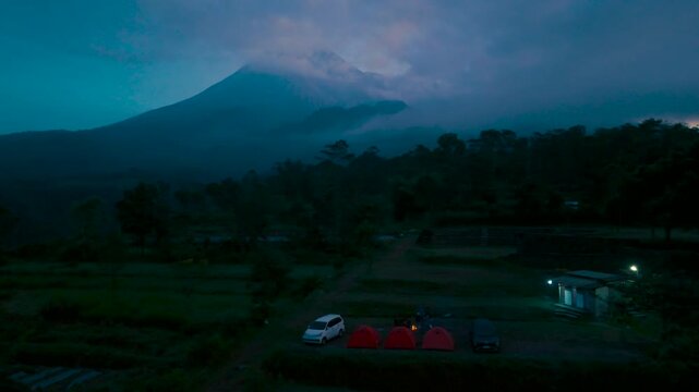 An aerial landscape showcasing a campsite with tents and vehicles, against a backdrop of a majestic mountain partially veiled by clouds during twilight, evoking a sense of adventure and tranquility.
