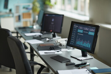 Row of empty office desks with computer monitors displaying data analytics dashboard, keyboards and mice arranged on workstations, modern workspace illuminated by natural light from large windows