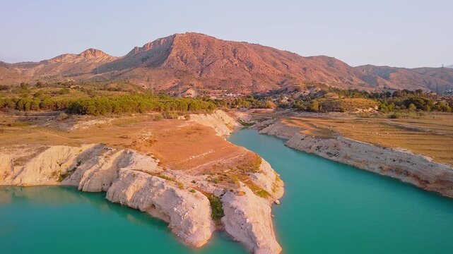 Drone video showing a sharp rocky peninsula surrounded by vibrant turquoise waters of a Spanish lake, bathed in warm golden hour sunlight.