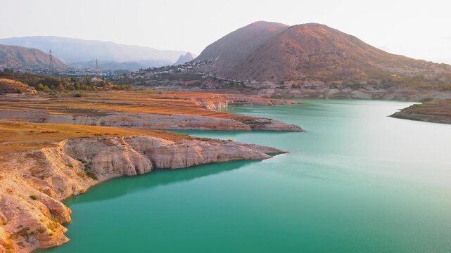 Drone video showing a sharp rocky peninsula surrounded by vibrant turquoise waters of a Spanish lake, bathed in warm golden hour sunlight.