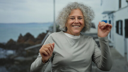 Elderly woman with grey hair joyfully dancing on a seaside promenade with a scenic ocean backdrop, capturing the essence of happiness and freedom in an outdoor setting.