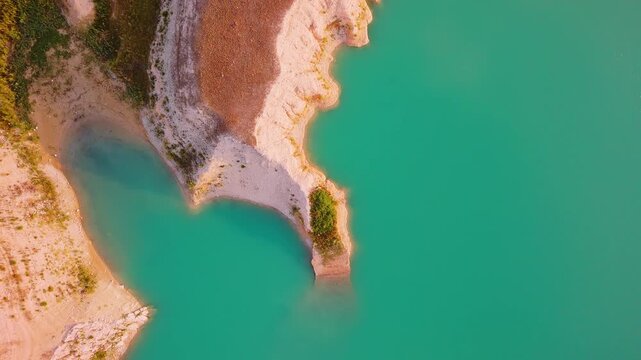 Drone video captures a rocky shoreline with green vegetation surrounded by bright turquoise waters of a lake in Spain, viewed directly from above.