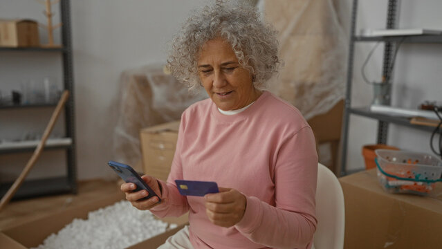 Woman using smartphone and credit card in new home living room unpacking cardboard boxes during move-in