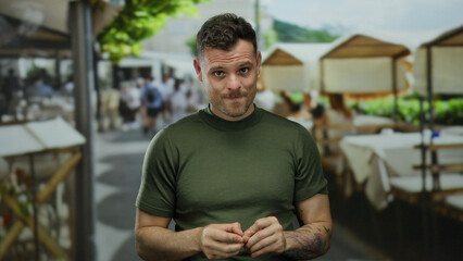 Young hispanic man in green shirt on urban terrace with tables and people, showcasing a distinctive tattoo, amidst a lively outdoor restaurant in a bustling city setting.