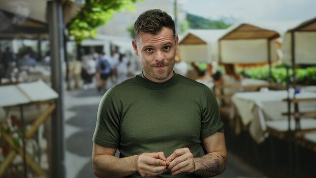 Young hispanic man in green shirt on urban terrace with tables and people, showcasing a distinctive tattoo, amidst a lively outdoor restaurant in a bustling city setting.