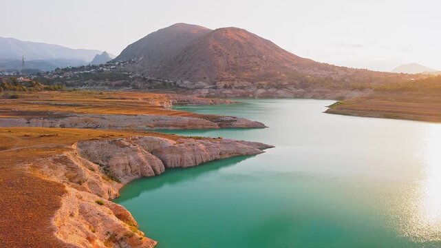 Drone video showing a sharp rocky peninsula surrounded by vibrant turquoise waters of a Spanish lake, bathed in warm golden hour sunlight.