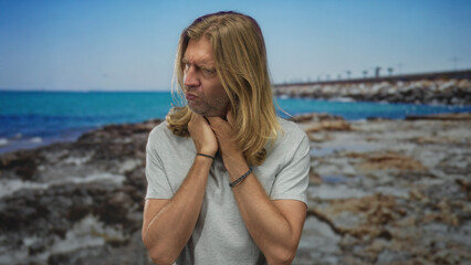 Blond man holds throat with both hands in pain on rocky coastline shore amid calm turquoise waves;...