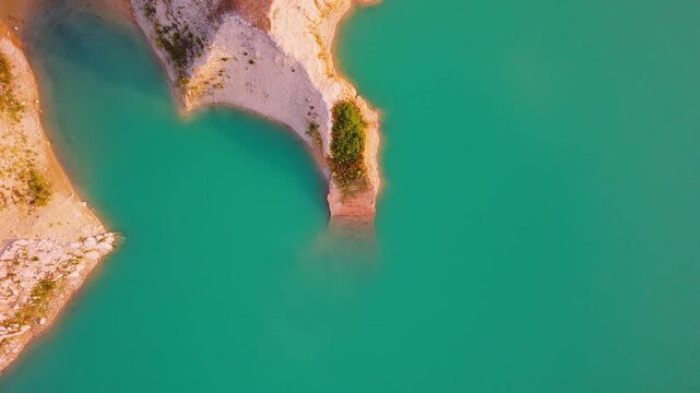 Drone video captures a rocky shoreline with green vegetation surrounded by bright turquoise waters of a lake in Spain, viewed directly from above.