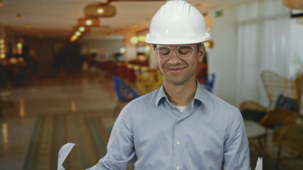Man holding plan in building lobby wearing hardhat and glasses with rolled documents in hand;...