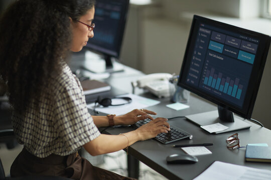 Young adult woman working at desk using desktop computer with data analytics dashboard on screen, typing on keyboard, sitting in modern office environment, focused on task