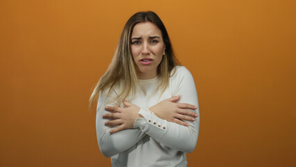 Obraz premium Young blonde woman feeling cold in a white sweater, hugging herself against an isolated orange background, expressing discomfort and shivering slightly.