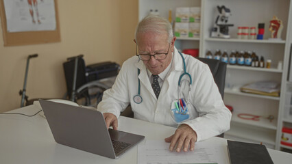 Senior male doctor in a clinic room working on a laptop with medical documents on the desk, surrounded by equipment and medicine shelves nearby.