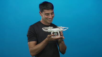 Young hispanic man holds a white drone in one hand and points finger to object in studio; curiosity.