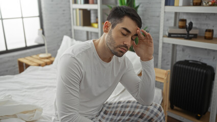 Obraz premium Young man sitting on bed in bedroom with tired expression wearing pajamas surrounded by modern furniture suggesting a relaxed home environment