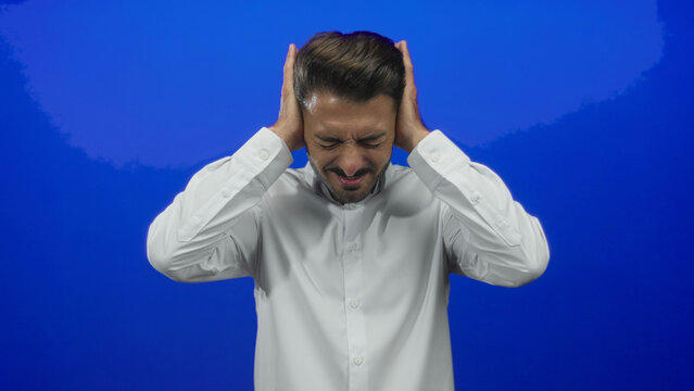 Young hispanic man wearing button down white shirt covers ears with hands and grimaces in blue studio shoot; pain.