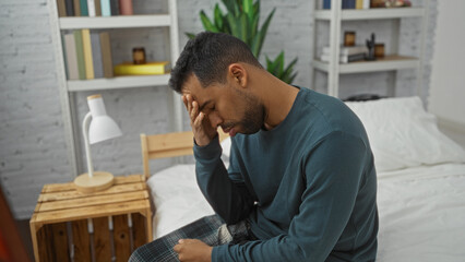 Obraz premium Young hispanic man sitting on bed with hand on forehead in cozy bedroom, displaying stress or fatigue, surrounded by books and plants suggesting a home interior setting.