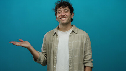 Hispanic man smiling against blue background with open hand gesture wearing beige shirt and white...