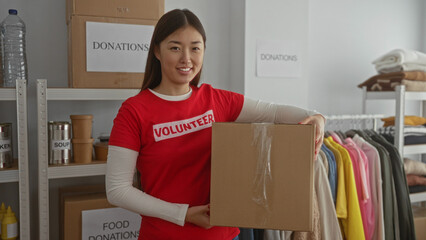 Woman in red shirt smiling holding box in charity donation center with shelves of food and clothes showcasing indoor volunteer work.