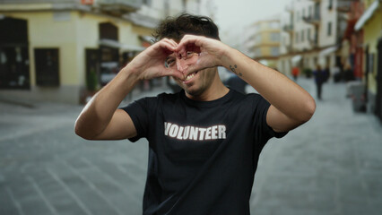 Hispanic man smiling outdoors wearing a volunteer shirt on a street, showcasing a cheerful expression against an urban backdrop, conveying themes of generosity and community spirit.
