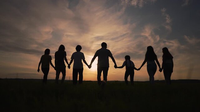 People hold hands during sunset creating silhouette of family and group together in open field showing unity and support with holding hand gesture and linked posture beneath warm clouded sky