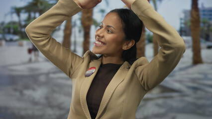 Hispanic woman smiling with arms raised wearing i voted badge outdoors by palm trees on street in united states.