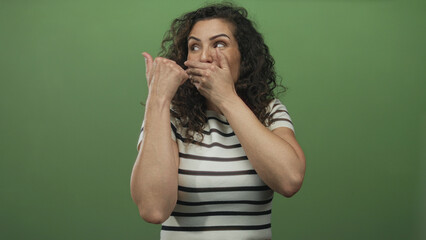 Young hispanic woman covers mouth with hand and points thumb to side in green studio; surprise reaction.