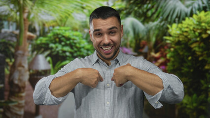 Young hispanic man in striped shirt with rolled sleeves points thumbs to chest in forest;...